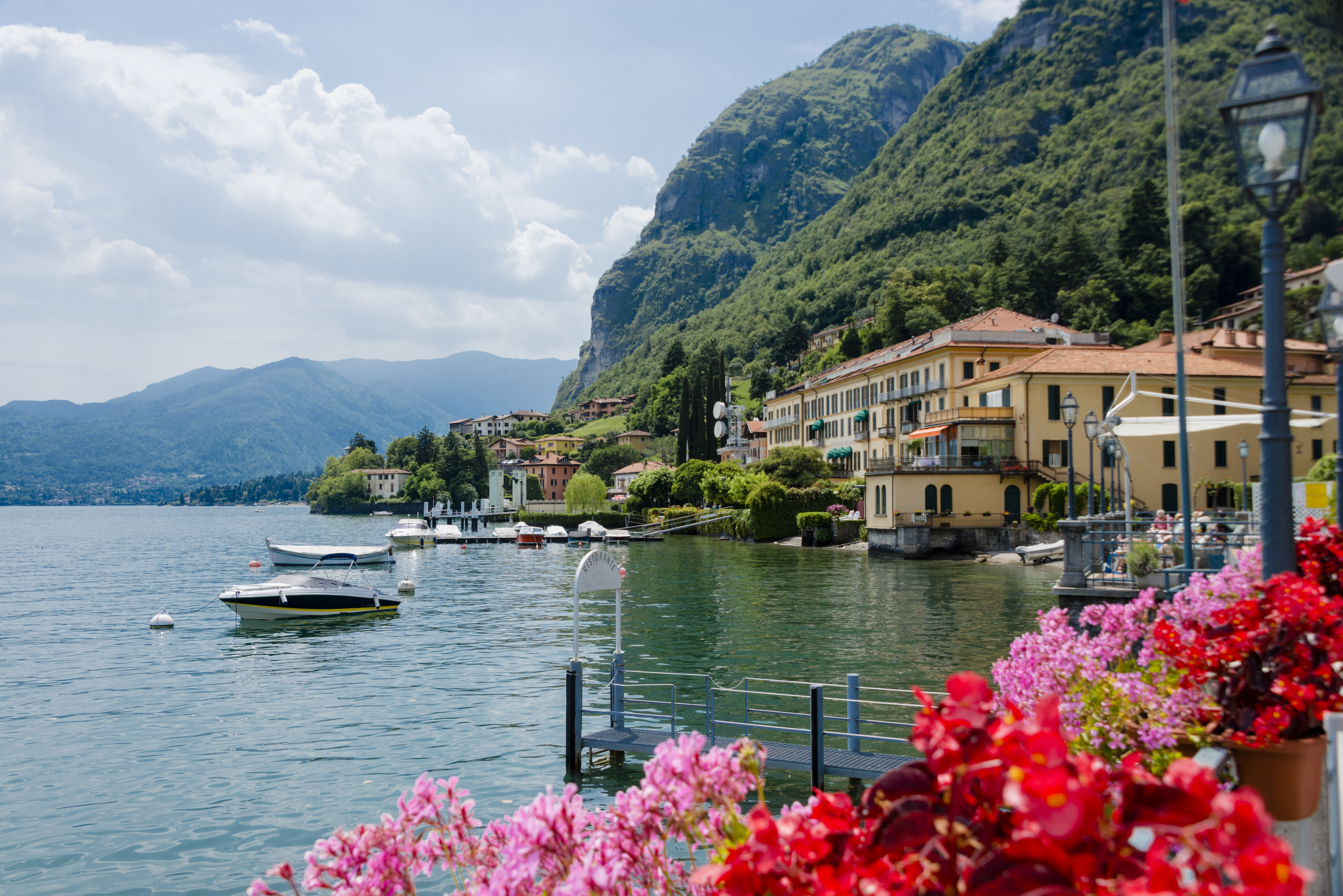 Red and pink geraniums on side of lake with small boats and the village of Menaggio on Lake Como