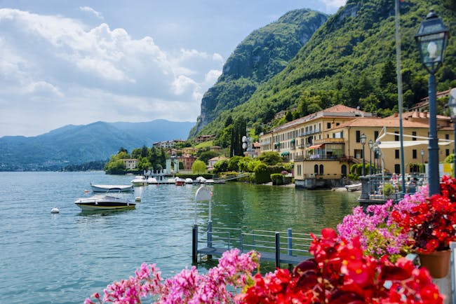 Red and pink geraniums on side of lake with small boats and the village of Menaggio on Lake Como