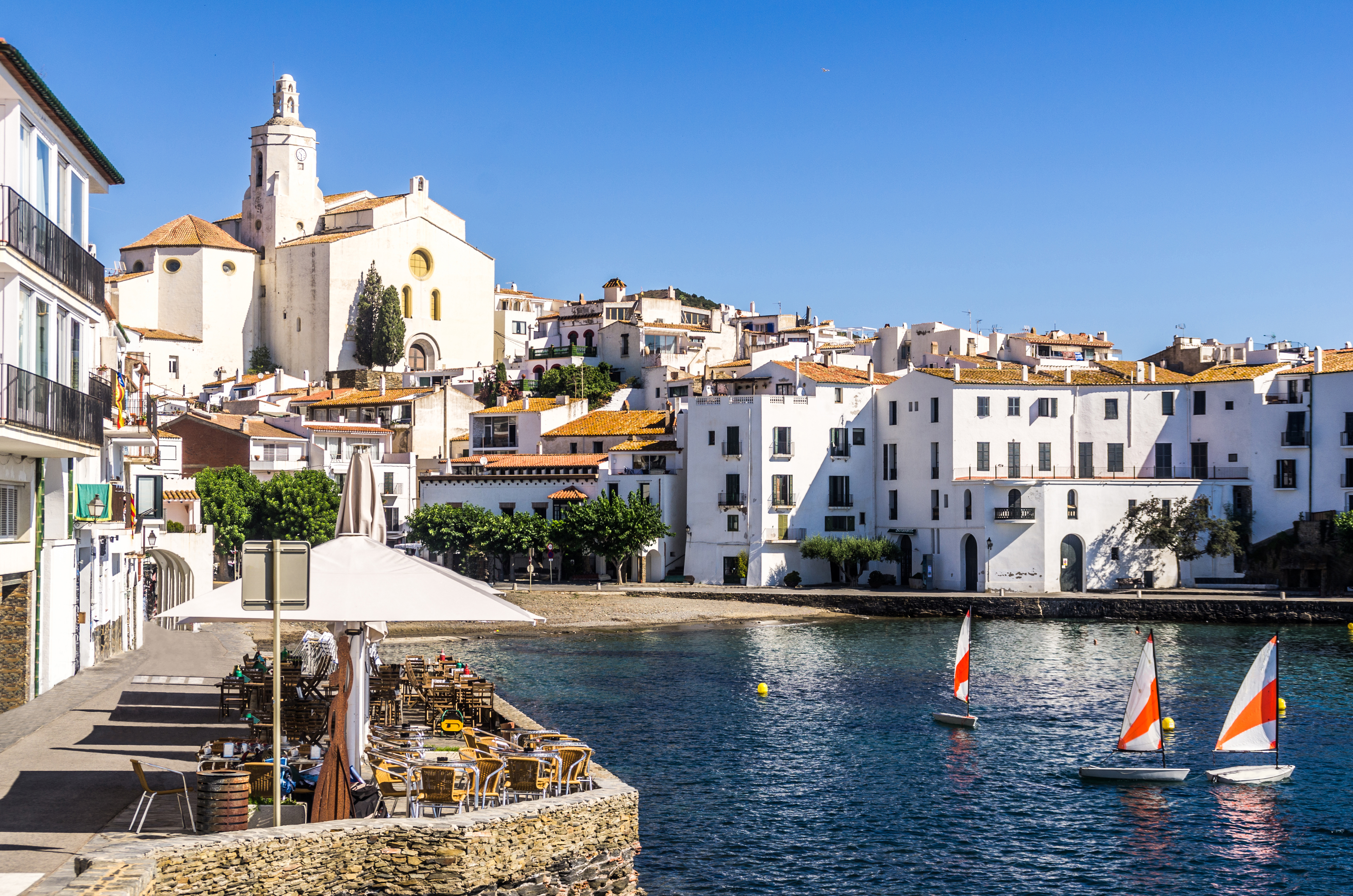 Harbour with small sail boats and white houses lining the seafront, cafe on one side, in Costa Brava town of Cadaques