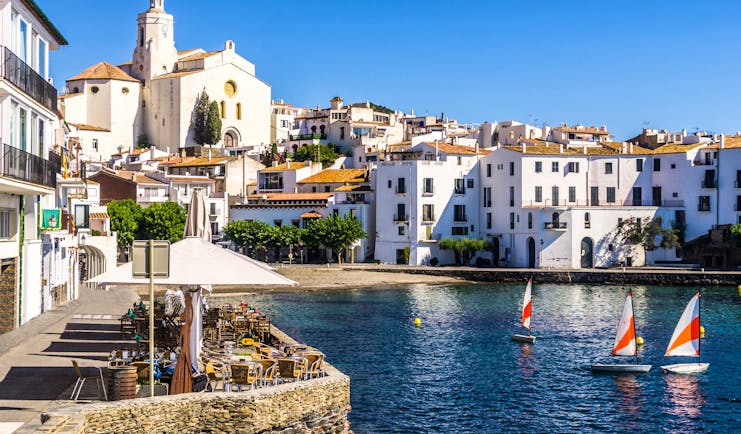 Harbour with small sail boats and white houses lining the seafront, cafe on one side, in Costa Brava town of Cadaques