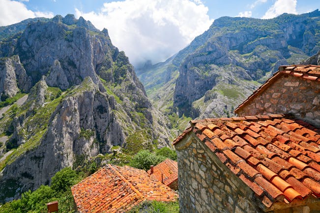 Rugged grey peaks with clouds and stone huts with red tiled roofs in foreground in the Picos de Europa